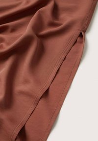 Un tissu couleur rouille tombe en doux plis, révélant une texture lisse et un éclat subtil. Il présente des coutures soignées le long des bords et une fente latérale pour un détail de design.