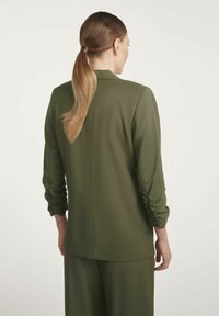 Kobieta z długim kucykiem, ubrana w oliwkową marynarkę i pasujące spodnie, stojąca tyłem na białym tle.