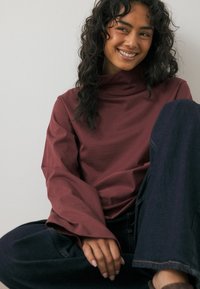 Mörk röd långärmad topp med hög krage, lös passform och vida ärmar, i kombination med mörka denimbyxor, på en modell som sitter mot en enkel vägg.