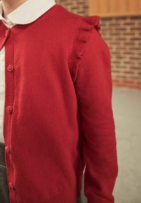 Cardigan rouge en tricot avec détail volanté sur les épaules, porté par-dessus une chemise blanche à col, sur fond de mur en briques flou.