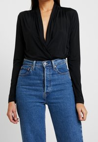 Schwarze Wickelbluse mit langen Ärmeln, tiefem V-Ausschnitt, aus weichem Stoff. Kombiniert mit hochgeschnittenen blauen Jeans aus Denim, die einen Knopfverschluss haben.
