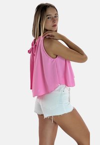 Różowy top bez rękawów o luźnym kroju, z marszczonym tyłem i kokardą, zestawiony z białymi, przetartymi szortami jeansowymi.