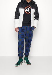 Svart och vit vadderad jacka med luva; svart hoodie med logotyp; rutiga mjukisbyxor i blått, grönt och rött; gröna och svarta höga sneakers.