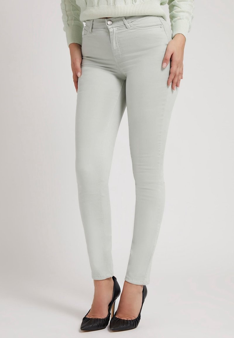 Kvinna iklädd ljusgrå skinny jeans med framfickor och svarta högklackade skor, stående mot en enfärgad bakgrund.