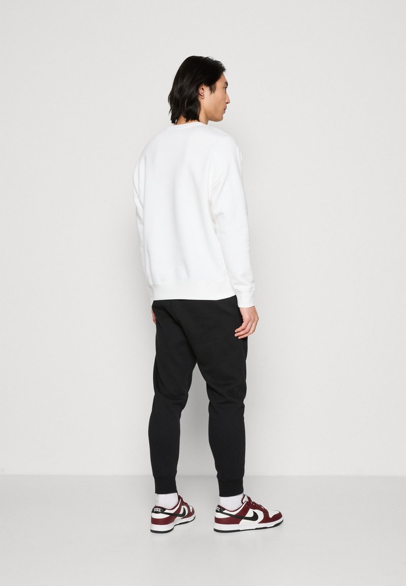 Sudadera blanca, pantalones jogger negros, con puños y cinturilla elásticos. Zapatillas rojas y blancas con un logo en blanco y negro. Fondo liso.