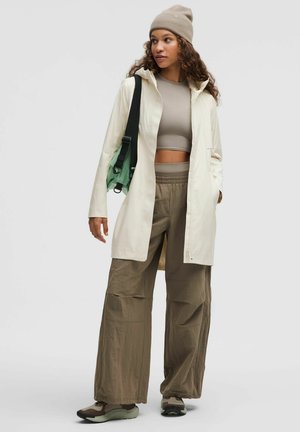 Lång vattentät jacka i krämfärg, beige kort topp, lösa byxor i olivgrönt, grå sneakers och en matchande mössa. Inkluderar axelväska med gröna detaljer.
