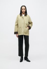 Lysebeige jakke med brune detaljer, der har en krave, to forlommer og en afslappet pasform. Kombineret med sorte jeans med lige ben og ankelstøvler.