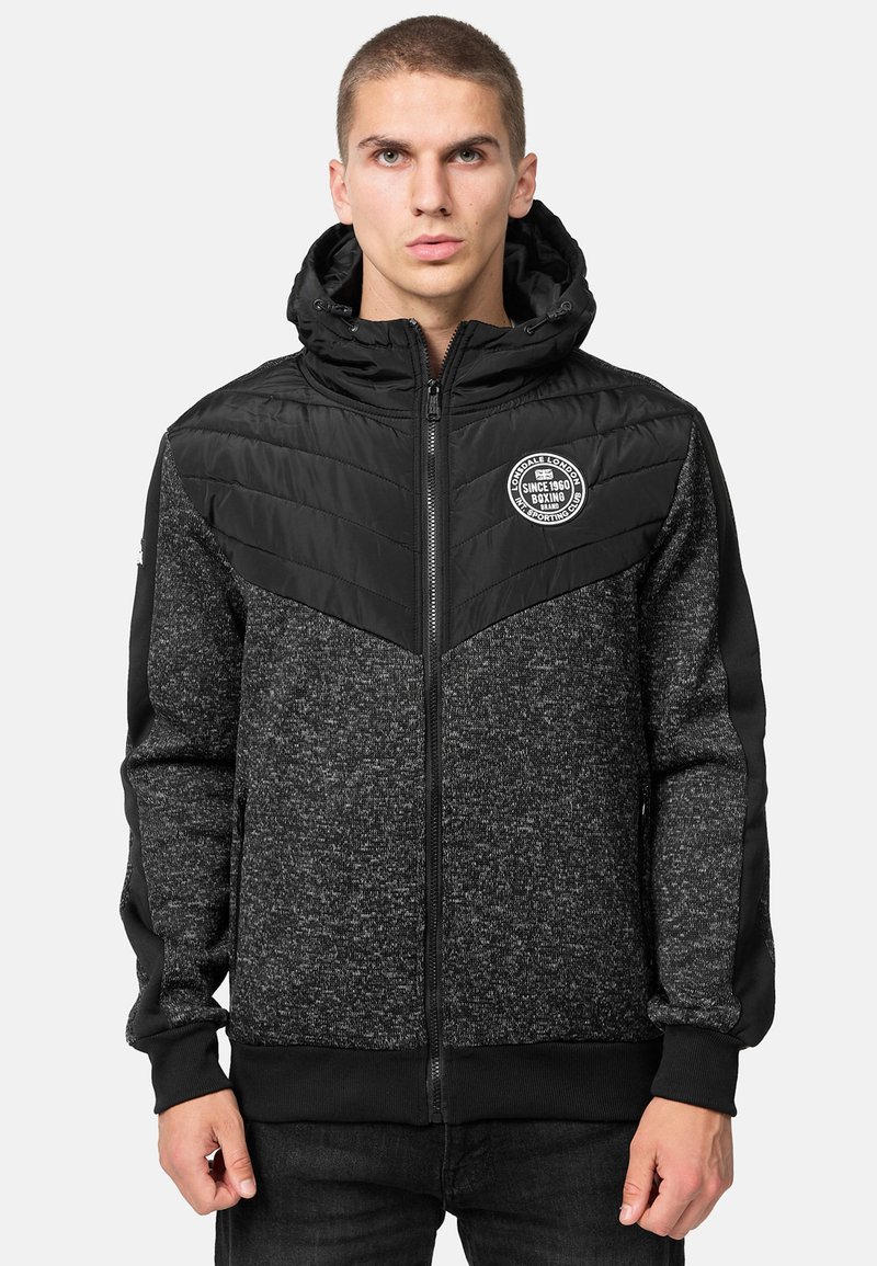 Zwarte en grijze zip-up jas met capuchon, voorzien van een gewatteerde bovenkant, elastische manchetten en een ronde logo op de borst.