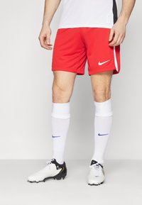 Rode sportshorts met het witte Nike-logo, gecombineerd met witte sokken met blauwe Nike-accenten en zwart-witte voetbalschoenen.
