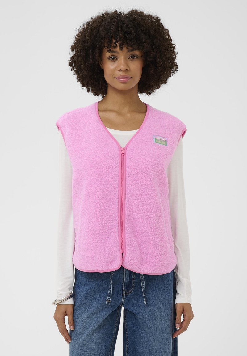 Frau mit lockigem Haar, die eine hellrosa, ärmellose Fleece-Weste mit Reißverschluss über einem weißen Langarmshirt und einer blauen Jeans trägt und aufrecht steht.