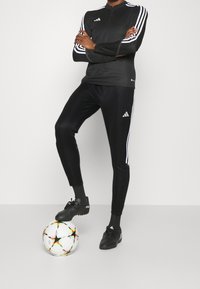 Černý sportovní top s dlouhým rukávem a zipem, spárovaný s černými zúženými kalhotami s bílými pruhy. Černé fotbalové boty a barevný míč.