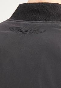 Svart bomberjacka med ribbad krage, med en broderad vingad emblem på baksidan. Slät textur med en minimalistisk design.