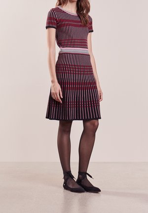 Kobieta w krótkim, paskowanym swetrowym sukienka w czerwieni, czerni i bieli, zestawiona z przejrzystymi czarnymi rajstopami oraz czarnymi balerinami z wiązaniami wokół kostek.