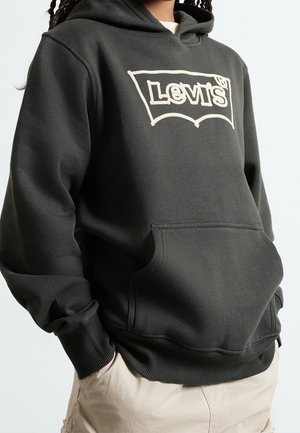 Persona vistiendo una sudadera con capucha negra de Levi's con logotipo bordado en blanco y pantalones beige, con las manos en el bolsillo de la sudadera, sobre un fondo claro y liso.