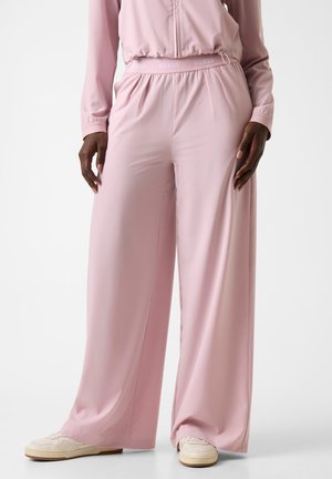 Persona che indossa pantaloni larghi a gamba larga color rosa pallido, a vita alta con elastico in vita, e scarpe da ginnastica bianche, in piedi contro uno sfondo bianco.