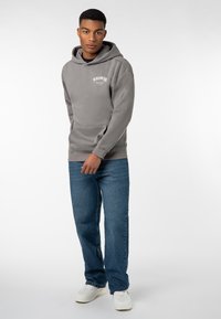 Grau-gefütterter Kapuzenpullover mit einer vorderen Tasche; verfügt über ein kleines weißes Logo. Kombiniert mit blauen Jeans und weißen Sneakern auf einem schlichten Hintergrund.