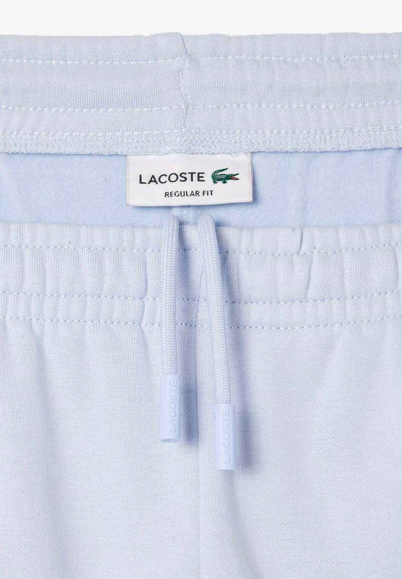 Pantalon de survêtement bleu clair avec une ceinture à cordon, arborant un label et un logo "LACOSTE". Tissu doux avec une coupe régulière.