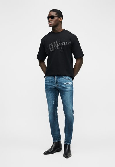 Άνδρας που φοράει μαύρα γυαλιά ηλίου, μαύρο T-shirt με λαμπερό κείμενο "Conique", μπλε σκισμένο στενό τζιν και μαύρα μυτερά μποτάκια, στέκεται με τα χέρια πίσω από την πλάτη.