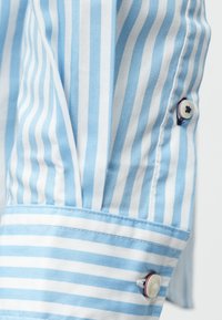 Tessuto della camicia a righe blu chiaro e bianche, con polsino a bottone caratterizzato da un motivo a stelle sul bottone. Texture liscia e design su misura.