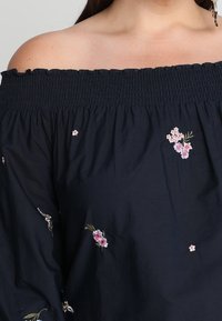 Femme portant un haut noir à épaules dénudées avec de petites broderies florales roses et des manches longues.