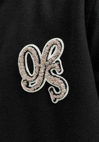 Il tessuto nero presenta un logo decorativo ricamato in una combinazione di beige e bianco, che mette in mostra lettere testurizzate con una finitura morbida.