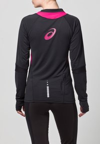 Donna che indossa un top atletico a maniche lunghe nero e rosa con fori per il pollice e una tasca con zip, mostrata di spalle su uno sfondo grigio.