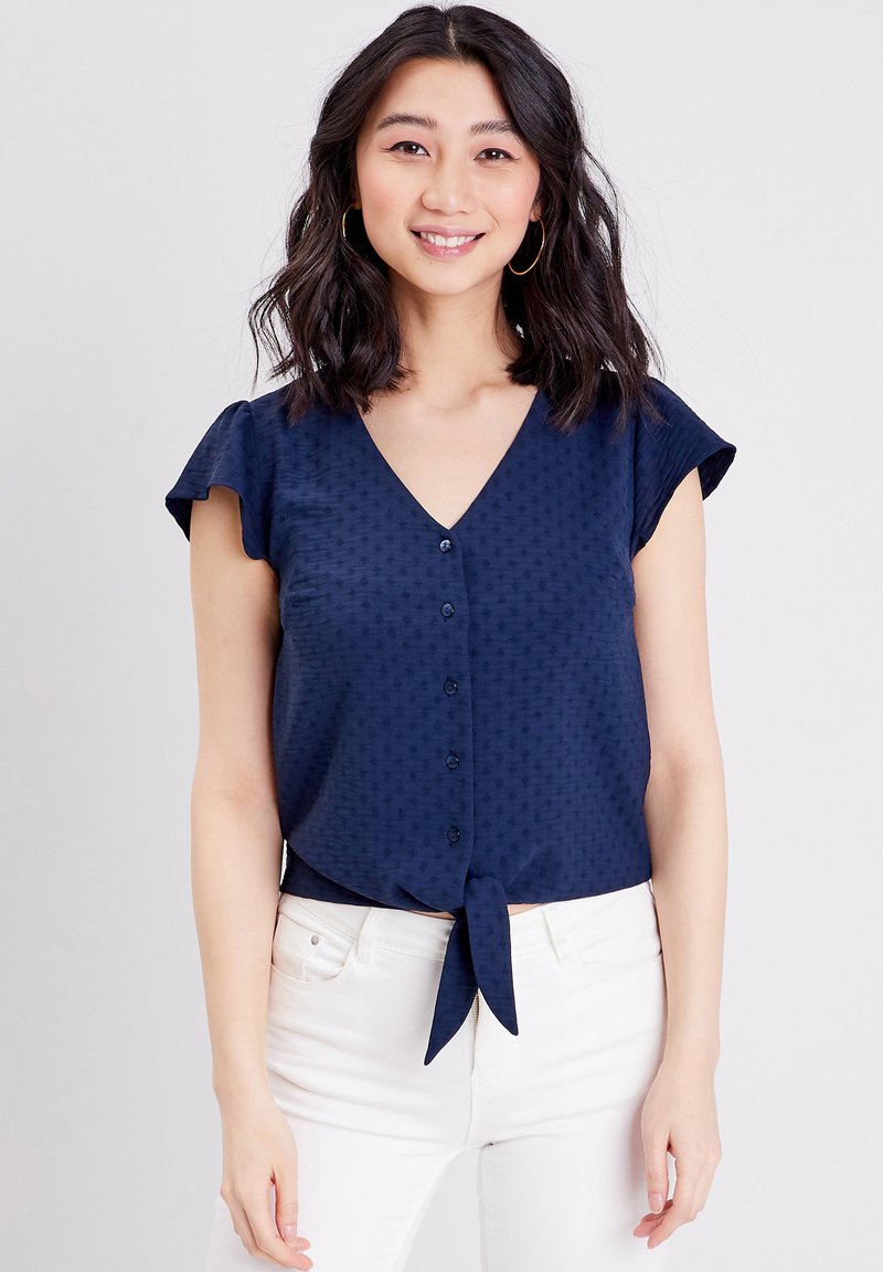 Blouse bleu marine à manches courtes avec un col en V, fermeture à boutons sur le devant, détail à nouer à la taille et motif texturé subtil sur toute la surface.