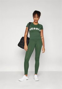 Zelený sportovní outfit s přiléhavým topem s krátkými rukávy označeným "NEBBIA" v bílé barvě a odpovídající legíny s vysokým pasem; doplněno černou sportovní taškou.