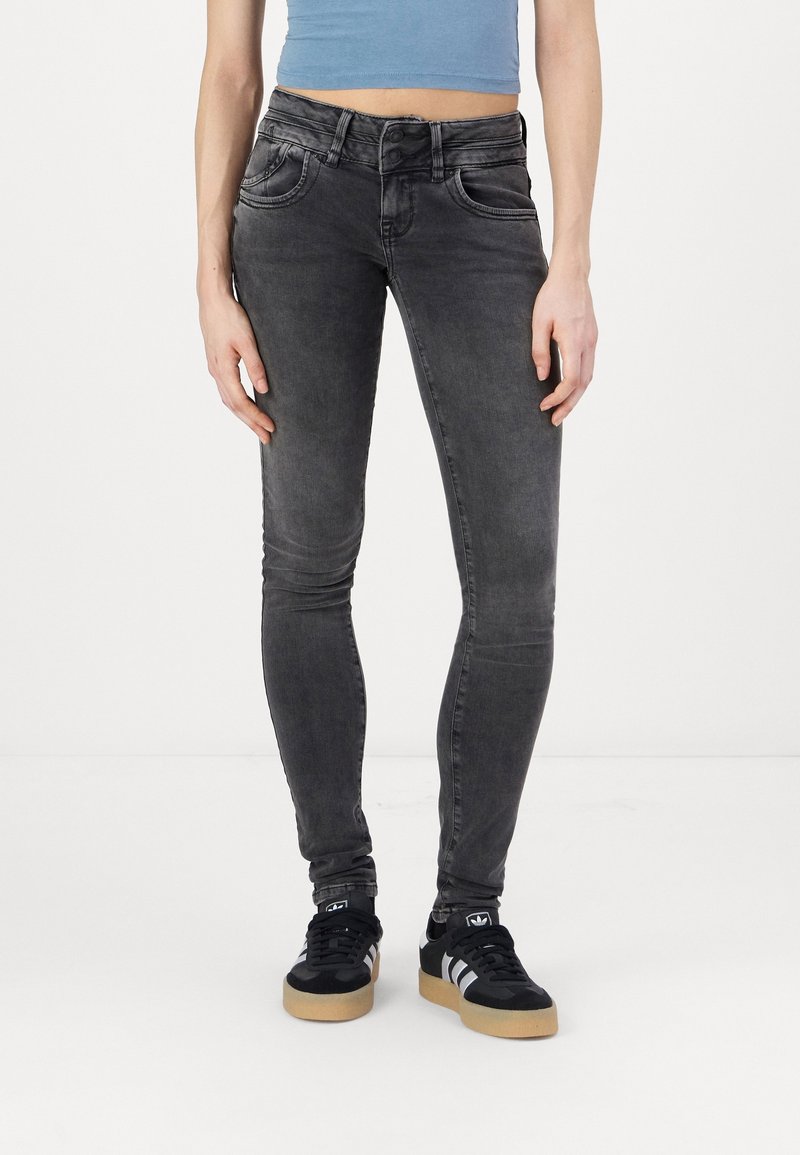 Jean noir slim porté par une personne debout, associé à des baskets noires et blanches et un top court bleu.
