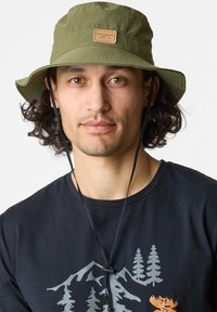 Cappello a secchielloo verde oliva realizzato in un materiale testurizzato e resistente all'acqua, con un patch in pelle sul davanti; dotato di una tracolla regolabile.