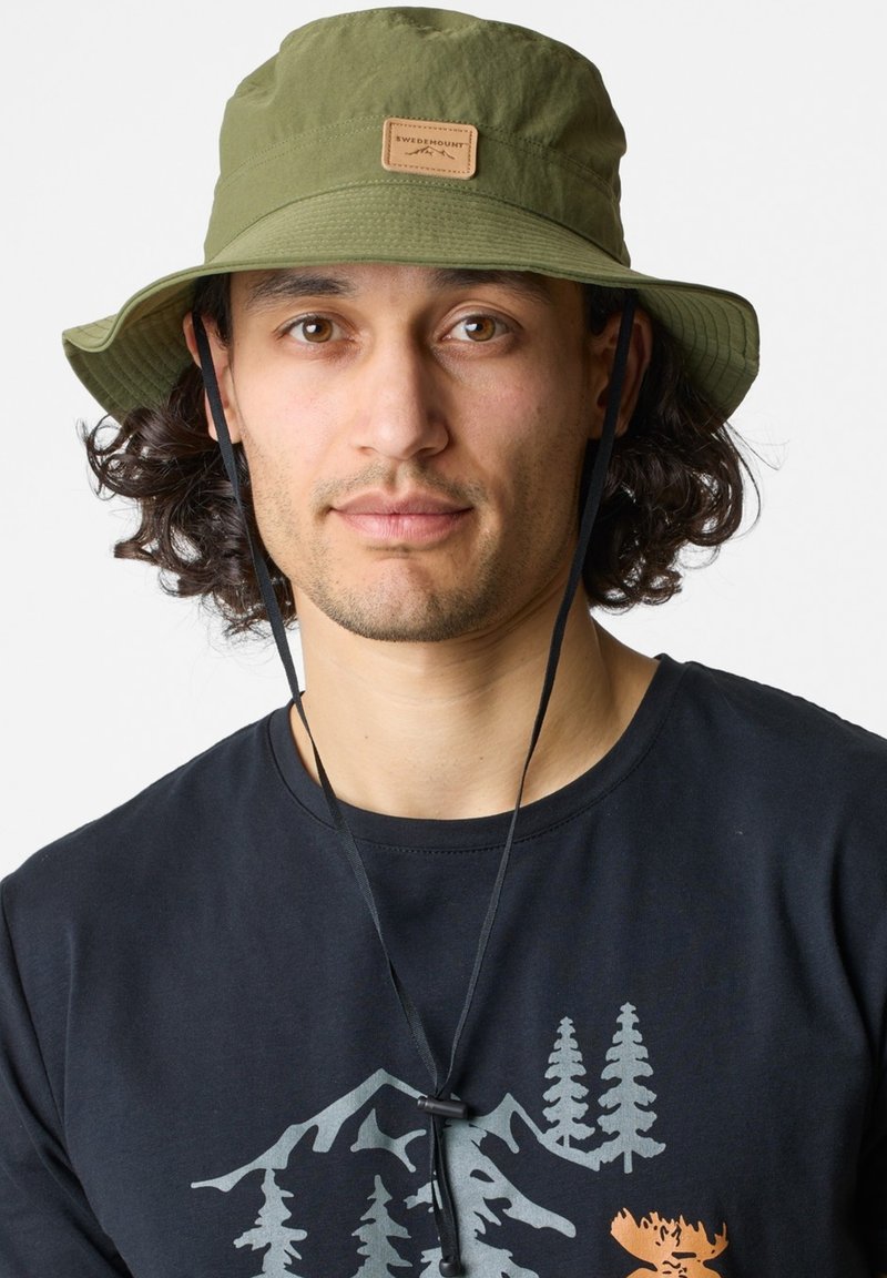 Cappello a secchielloo verde oliva realizzato in un materiale testurizzato e resistente all'acqua, con un patch in pelle sul davanti; dotato di una tracolla regolabile.
