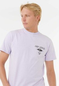 Lila Baumwoll-T-Shirt mit Rundhalsausschnitt, kurzen Ärmeln, Grafik auf der Vorderseite mit "RIP CURL"-Text und Designelementen. Sanfte Textur.