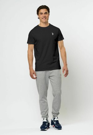 T-shirt nera in cotone con collo rotondo e piccolo logo bianco sul lato sinistro del petto, abbinata a pantaloni jogging grigi chiari e sneakers blu navy.