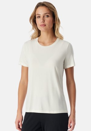 Vrouw met lichtbruin haar draagt een effen wit T-shirt met korte mouwen en een zwarte broek, staand tegen een effen lichte achtergrond.