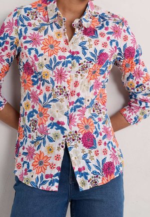 Personne portant une chemise blanche à manches longues avec des motifs floraux colorés et un jean bleu, debout les mains sur les hanches.