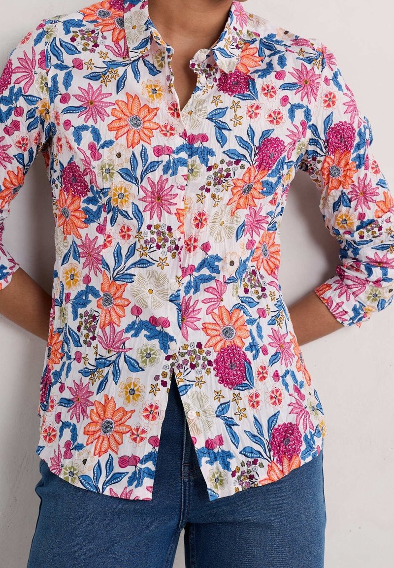 Personne portant une chemise blanche à manches longues avec des motifs floraux colorés et un jean bleu, debout les mains sur les hanches.