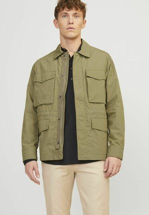 Veste utilitaire olive avec une fermeture à bouton, quatre poches avant et une coupe décontractée. Associée à une chemise noire et un pantalon clair.