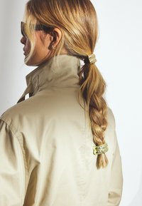 Gai beigtas trīskāršais mētelis uz sievietes ar austiem matiem, kas ir piestiprināti ar spīdīgu zelta matu aksesuāru. Mati ir gari un viļņaini.