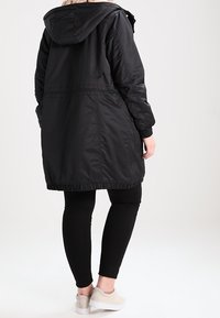 Černá parka dlouhé linie s kapucí, se staženým pasem a elastickým lemem. Hladký materiál s minimálním zdobením, nošená přes černé legíny.