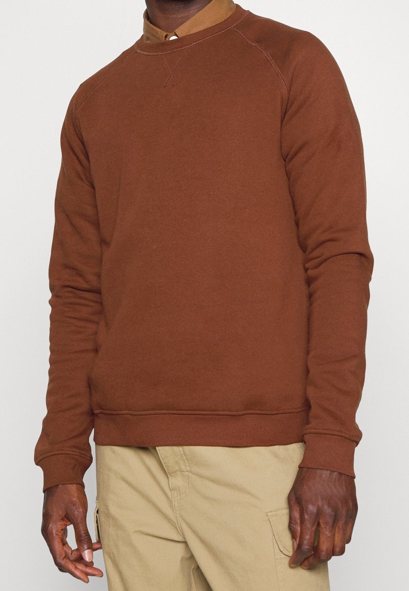 Brun bomullssweater med rund hals, ribbade ärmslut och nederkant. Har raglanärmar och en slät textur, bärs över en ljusbrun skjorta med krage.