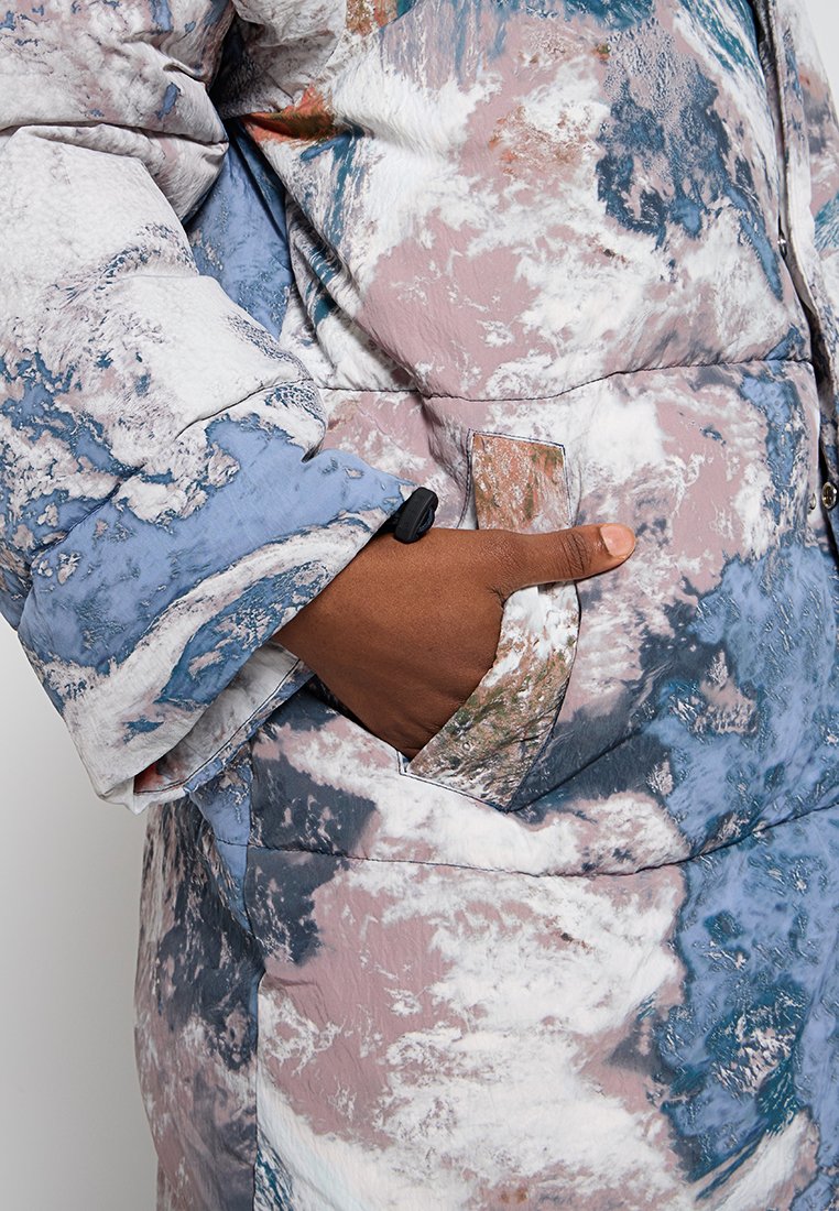 Manteau rembourré avec un motif abstrait bleu, rose et blanc. Il dispose d'une poche latérale et d'un tissu texturé et brillant.