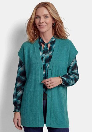 Donna con capelli rossi fino alle spalle che indossa un gilet lavorato a maglia azzurro-verde sopra una camicetta fantasia, sorridente su uno sfondo neutro.