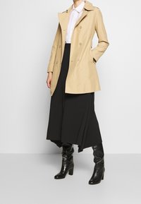 Gabardina doble abotonadura de color beige con cuello, combinada con una falda negra fluida hasta el tobillo y botas negras de tacón hasta la rodilla.