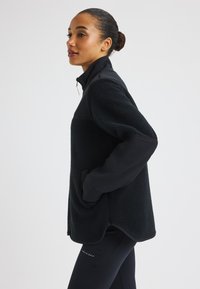 Zwarte fleec jacket met een gladde bovenstof, hoge kraag, zijzakken en een licht losse pasvorm. Getextureerd fleece lichaam met contrasterende mouwen.