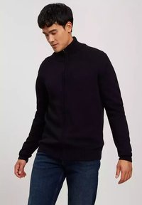 Czarny sweter na zamek z wysokim kołnierzem, z prążkowanymi mankietami i dolnym wykończeniem. Noszony z niebieskimi dżinsami, posiadającymi dopasowany krój i gładką teksturę.