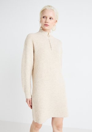 Robe en tricot beige avec un col montant, dotée d'une fermeture éclair et de manches longues. La texture est côtelée, avec une coupe légèrement décontractée.