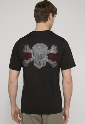 Mężczyzna noszący czarną koszulkę z dużym wzorem czaszki i skrzyżowanych kości z kryształkami na plecach, częściowo widoczne czerwone litery z kryształków.