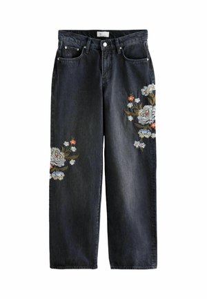 Kobieta w brązowym golfie włożonym do czarnych szerokich dżinsów z kwiatową haftem, czarnym paskiem i czarnych, spiczastych butach.