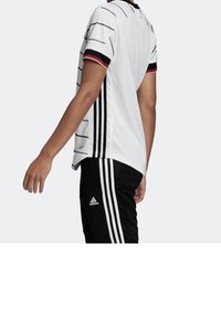 Valkoinen urheilu paita, jossa on mustia ja punaisia raitoja, yhdistettynä mustiin housuihin, joissa on valkoiset raidat ja Adidas-logo lahkeessa.