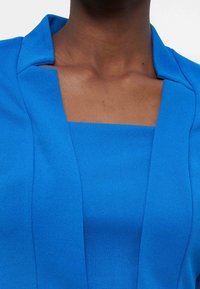 Gros plan sur une personne portant un blazer bleu vif et un haut à encolure carrée assorti sur un fond uni.